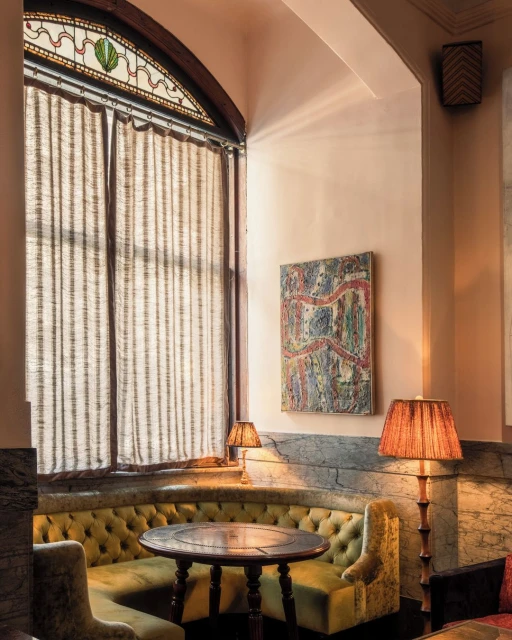 Cozy corner seating area with yellow-green velvet booth, round dark wood table, and patterned lamps in an elegant, vintage-style interior.