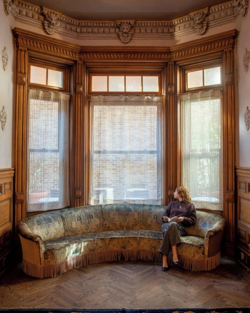 Adult woman seated on vintage curved couch inside of an ornate room with paneled walls and large windows overlooking the outdoors.