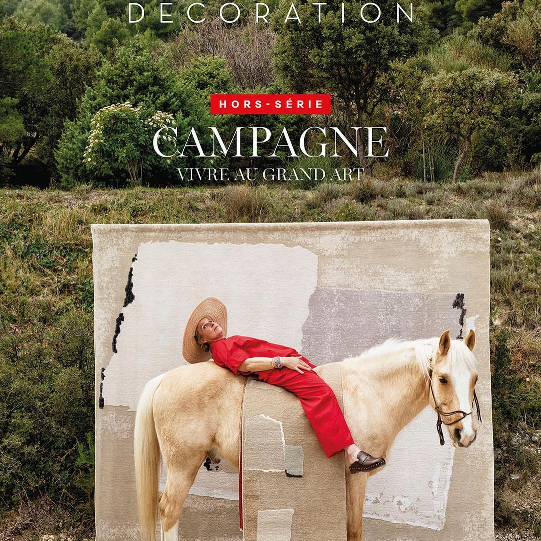 Person in red outfit reclines on a horse draped with textured fabric against a green, hilly landscape, as featured on a magazine cover.