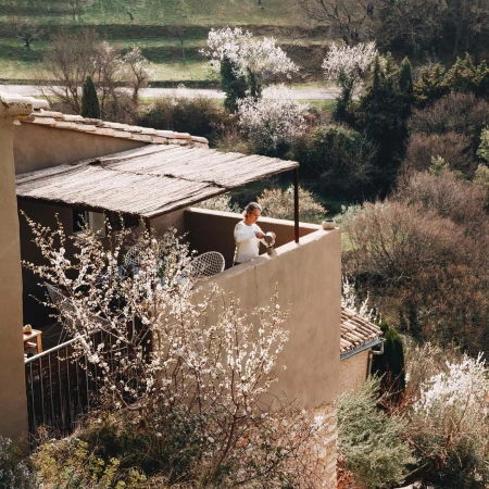 Adult female tends plants in pots on sunlit balcony. Plants in bloom surround rural house overlooking hillside with trees in background.