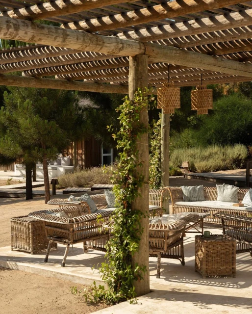 Outdoor patio area with wicker furniture set under a wooden pergola, with climbing vines and hanging wicker lamps. Natural light.