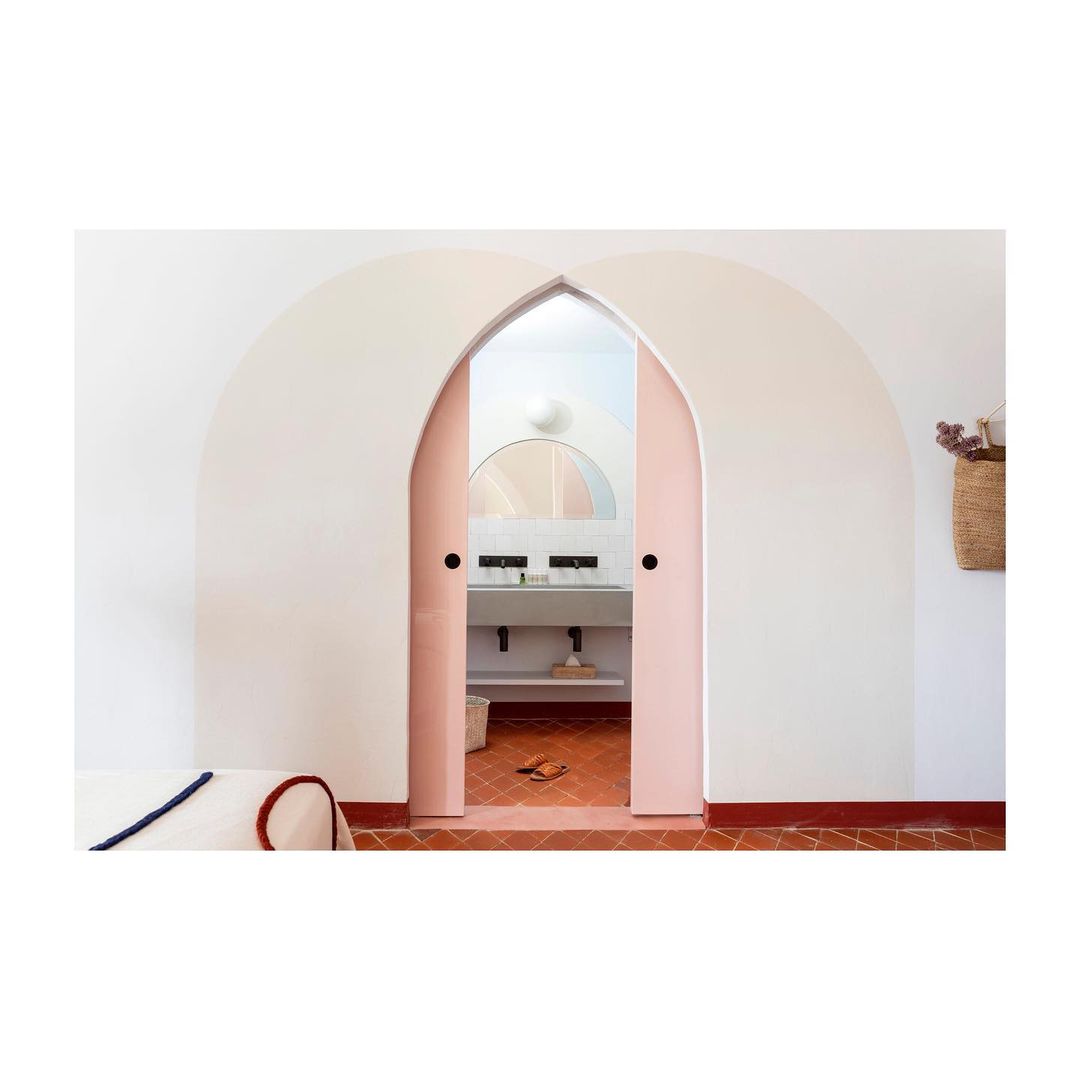 Arched doorway with partially open pink double doors revealing a bathroom with a sink, mirror, terracotta floor tiles and white walls.