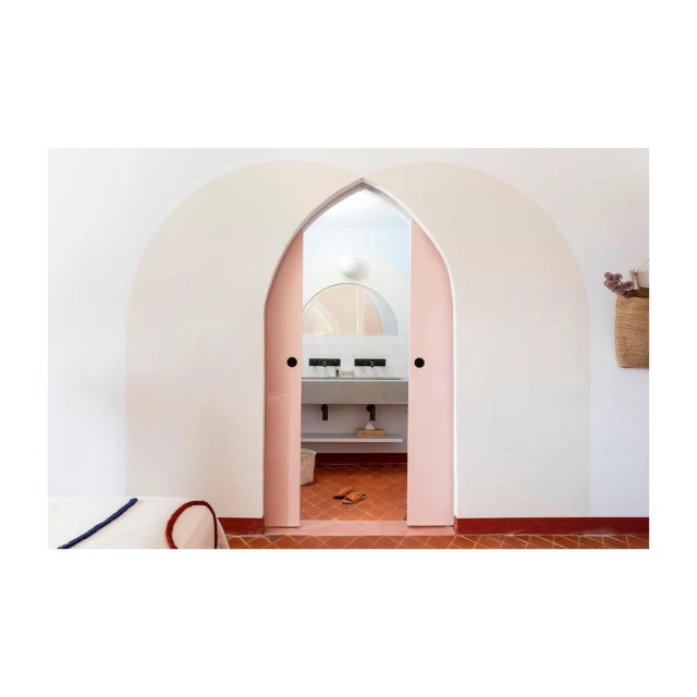 Arched doorway with partially open pink double doors revealing a bathroom with a sink, mirror, terracotta floor tiles and white walls.