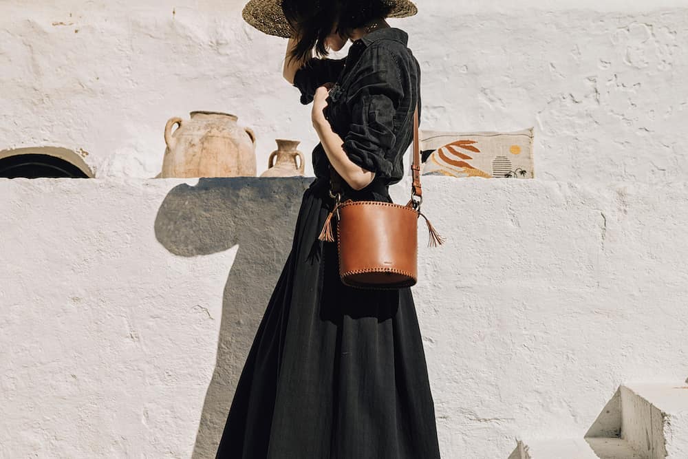 Woman in flowing black dress with brown bucket bag standing against white wall with decor, basking in sunlit outdoor scene.