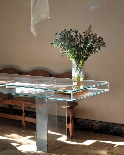 Still-life photograph of a transparent glass table topped with a floral arrangement indoors with natural light in a home interior setting.
