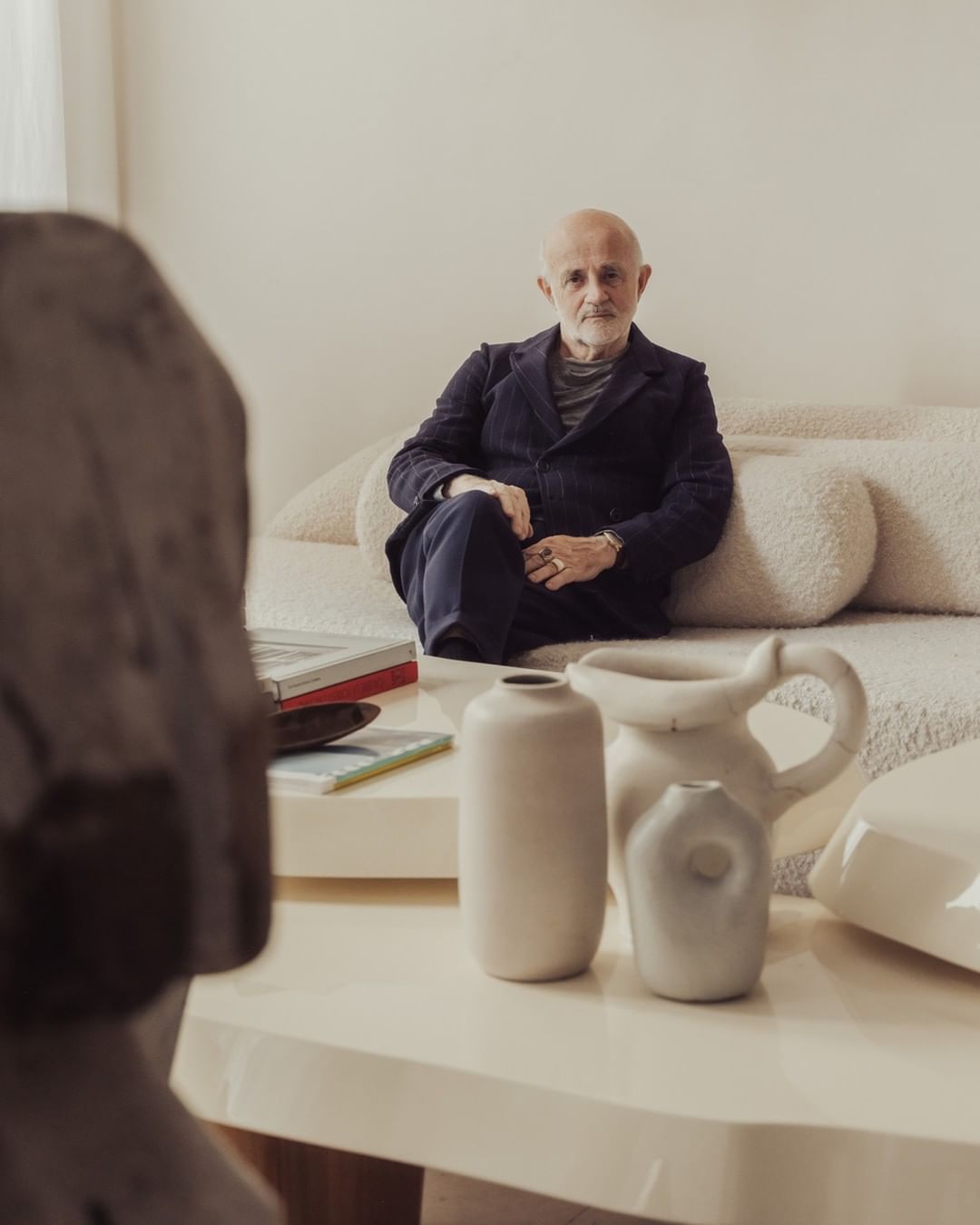 Mature man in a dark suit poses indoors on a cream couch near modern art objects and off-white walls in soft, natural lighting.