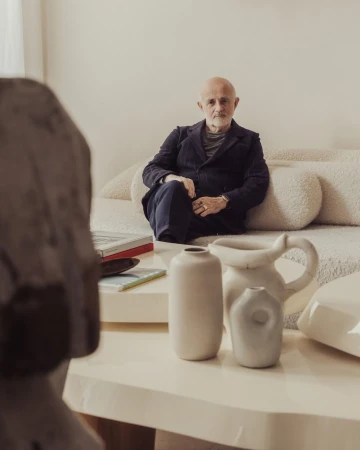 Mature man in a dark suit poses indoors on a cream couch near modern art objects and off-white walls in soft, natural lighting.