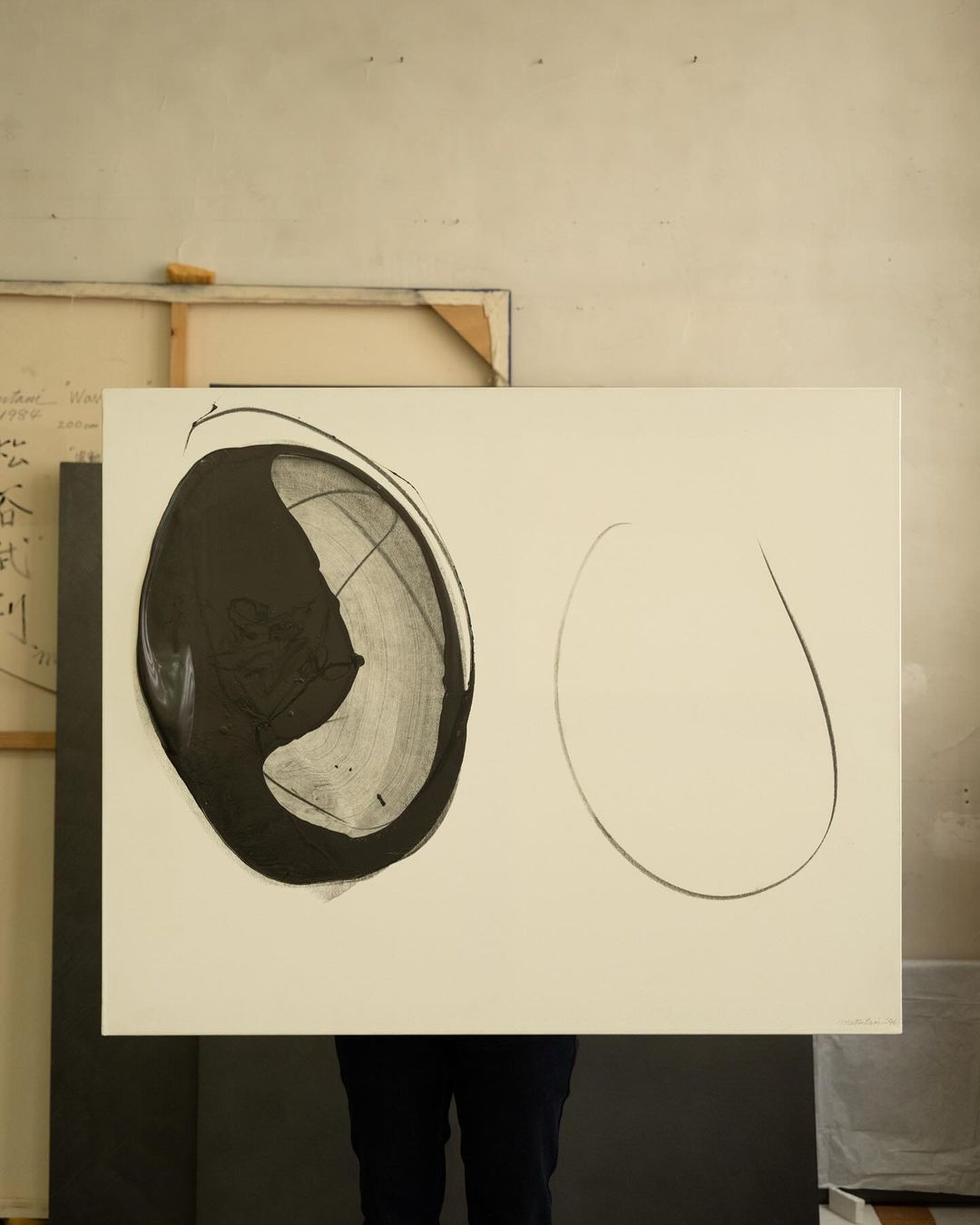 Abstract art featuring black and white shapes on a rectangular canvas, held by an unseen person against a muted beige studio backdrop.