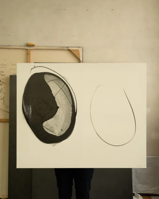 Abstract art featuring black and white shapes on a rectangular canvas, held by an unseen person against a muted beige studio backdrop.