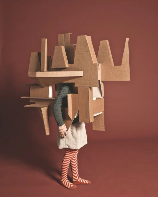 Person in cardboard box costume with gray sweater, patterned leggings, and red flats against a solid terracotta-red background in studio lighting.