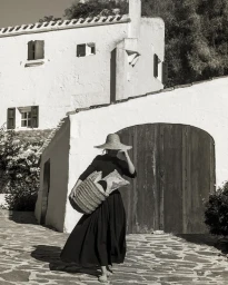 Woman wearing a wide-brimmed straw hat and black dress holding a woven basket outside a white stucco building.