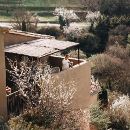 Person on a sunlit balcony pouring liquid into a cup against a natural landscape background with blooming trees.
