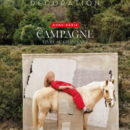 Woman in red jumpsuit and straw hat lying across the back of a light-coloured horse outdoors by a patterned rug.
