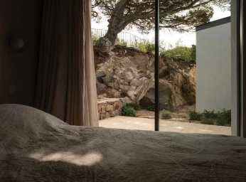 Interior view from a bed looking through a large glass sliding door at a rocky hillside and mature tree in Paris by Karel Balas
