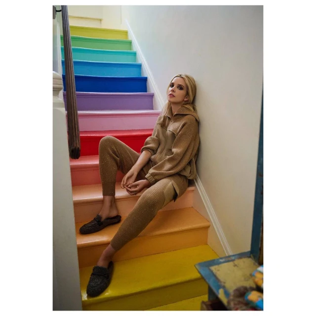 Adult woman in knitwear loungewear sits on rainbow painted stairs indoors, casual fashion concept, neutral light, lifestyle editorial.