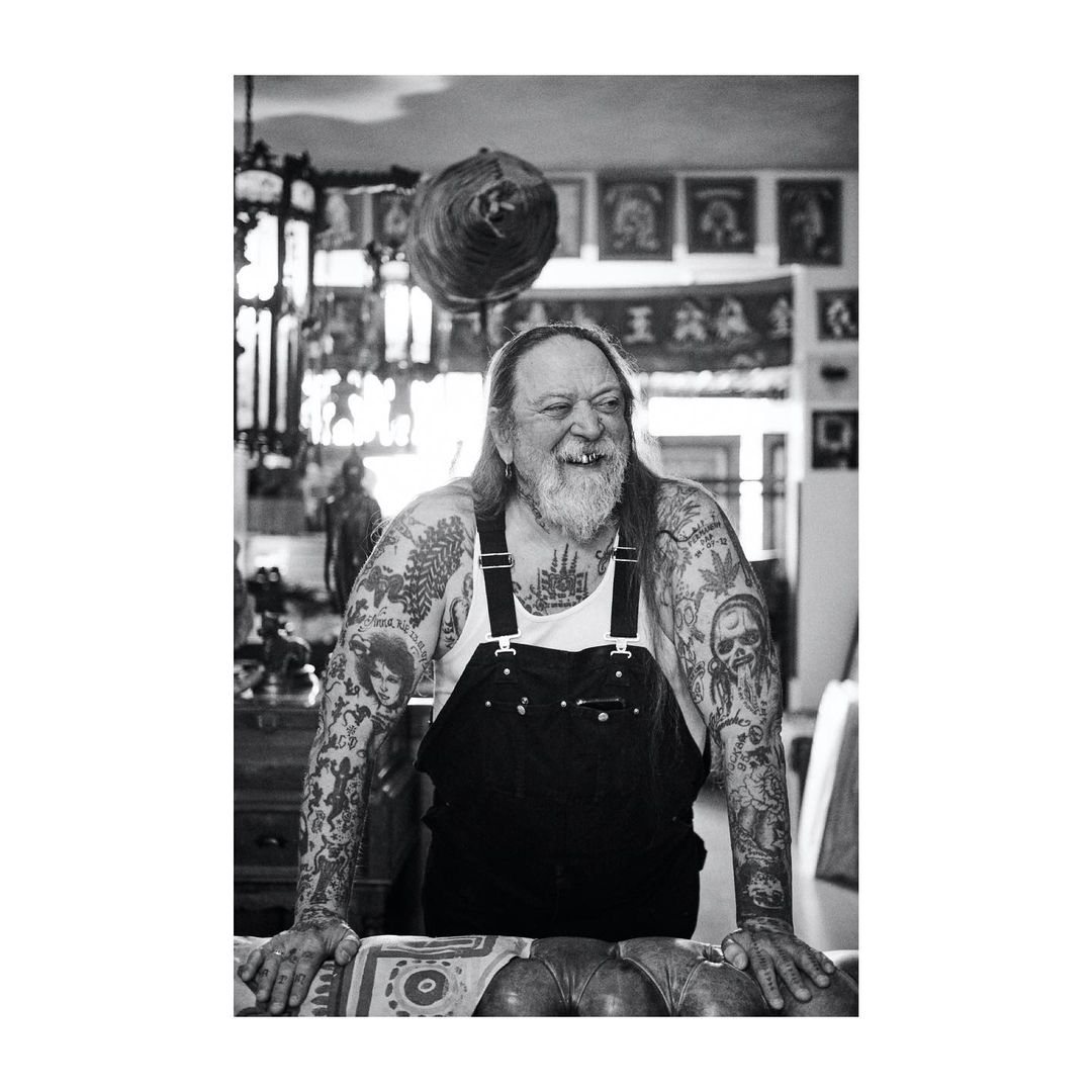 Monochrome portrait of a tattooed, older adult man with a beard and overalls, leaning on a patterned table in an indoor setting.