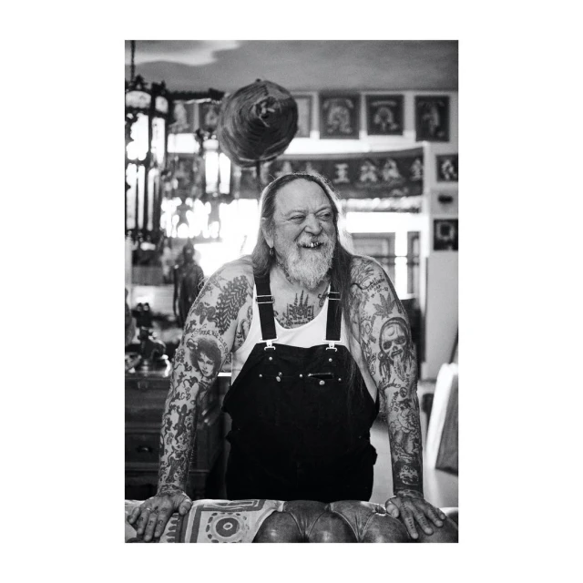 Monochrome portrait of a tattooed, older adult man with a beard and overalls, leaning on a patterned table in an indoor setting.