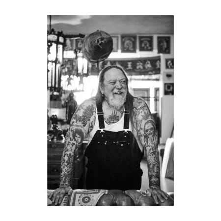 Monochrome portrait of a tattooed, older adult man with a beard and overalls, leaning on a patterned table in an indoor setting.