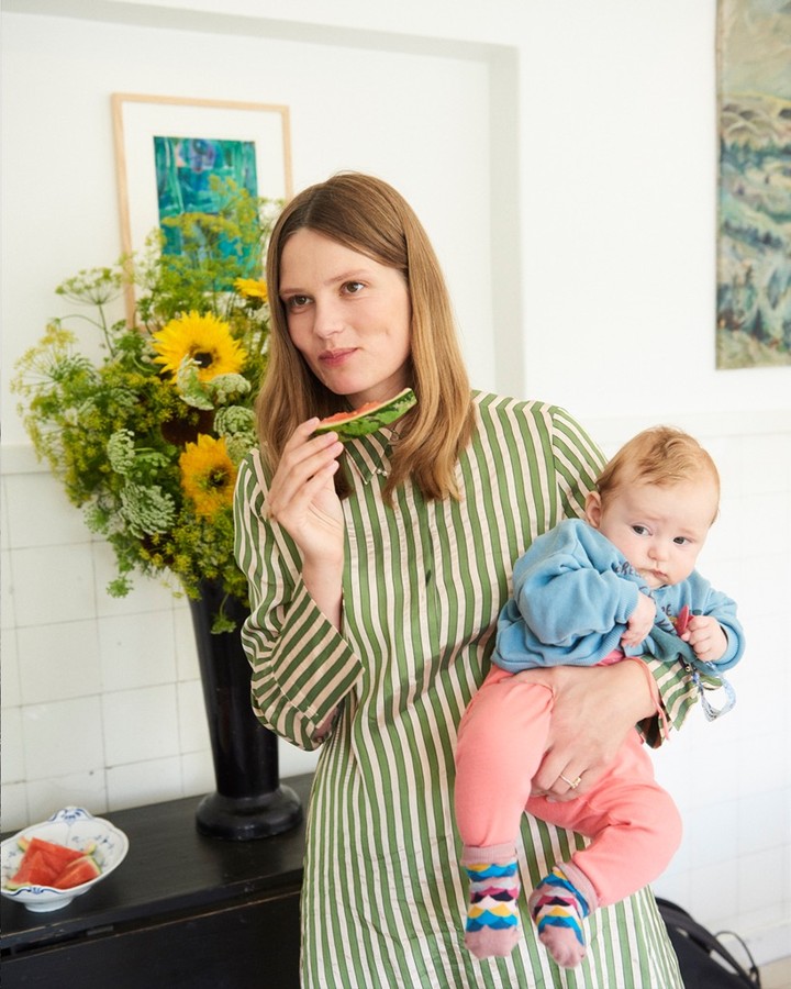 Woman and baby indoors with sunflowers and paintings. Woman eating watermelon in striped shirt, holding baby with patterned socks.