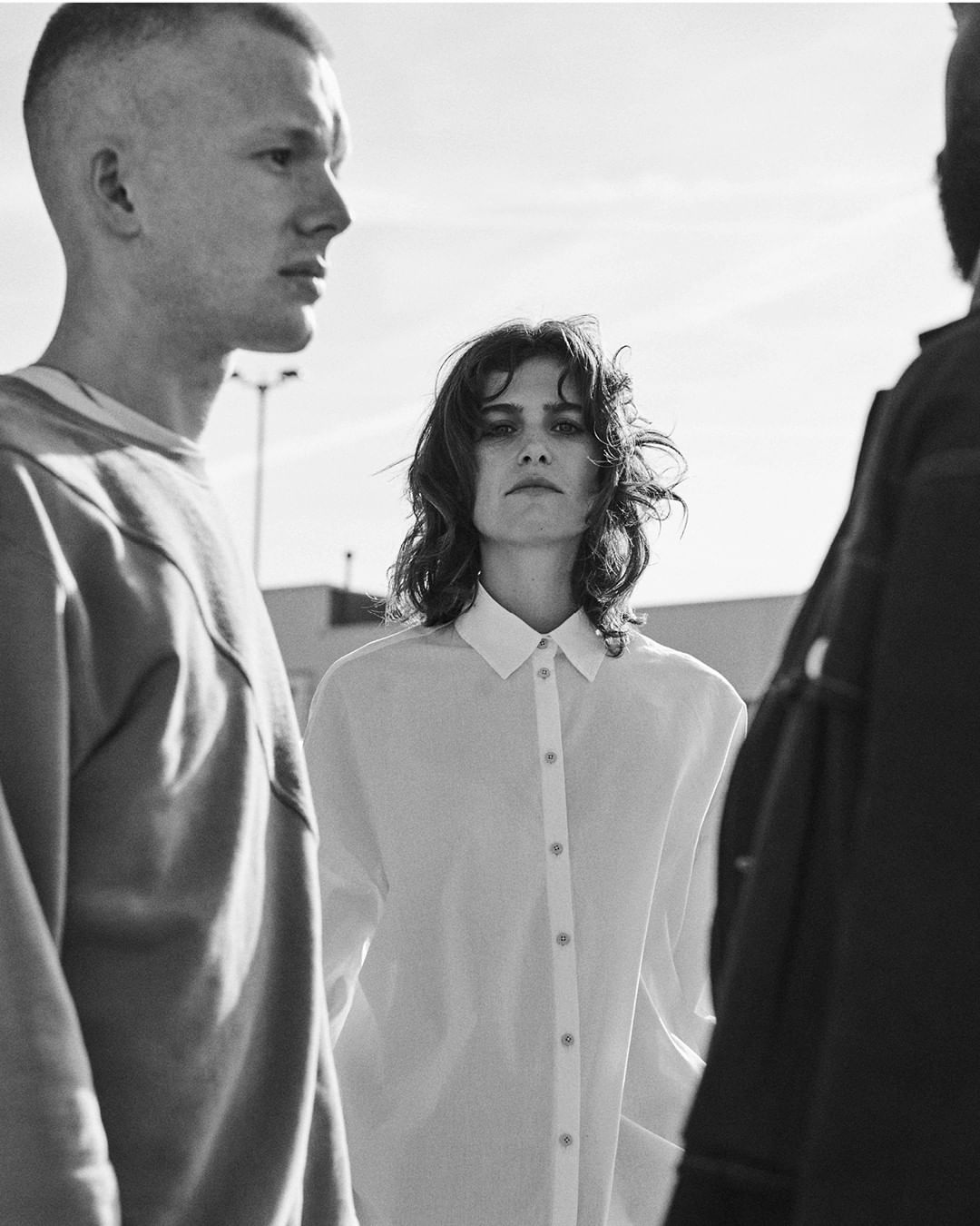 Black and white studio shot of a woman in a button-down shirt, flanked by two adult males on an outdoor urban rooftop.