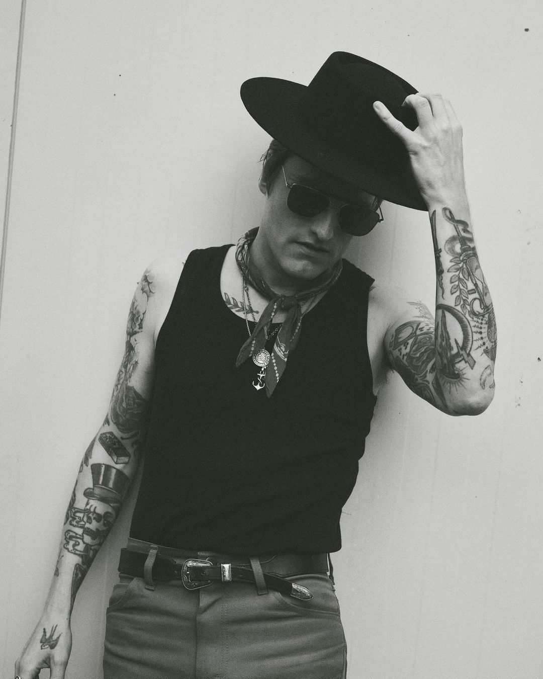 Monochrome portrait of a tattooed man wearing a black hat and sunglasses, posing against a light gray wall outdoors.