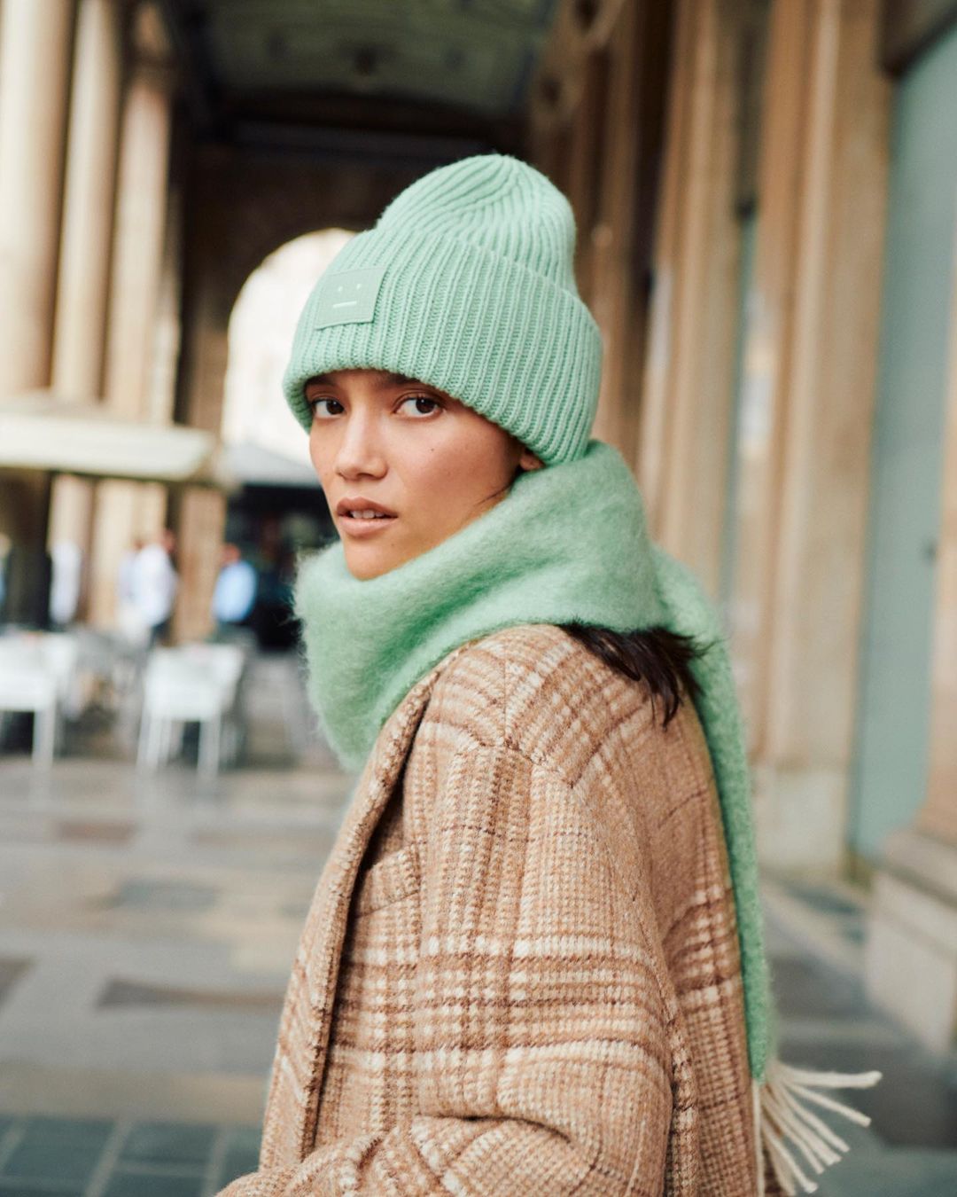 A woman in a beanie and scarf poses outdoors with stone columns. She is wearing a plaid coat in neutral tones and looking at the camera.