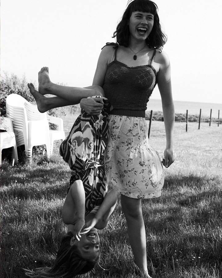 Black and white shot of a woman holding a smiling child upside down in a grassy field on a sunny day, with a fence in the background.