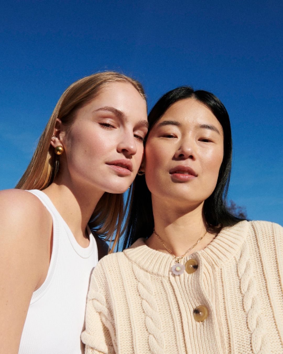 Two women pose outdoors against a clear blue sky. The woman on the left has blonde hair and wears a white tank top, while the other has dark hair.