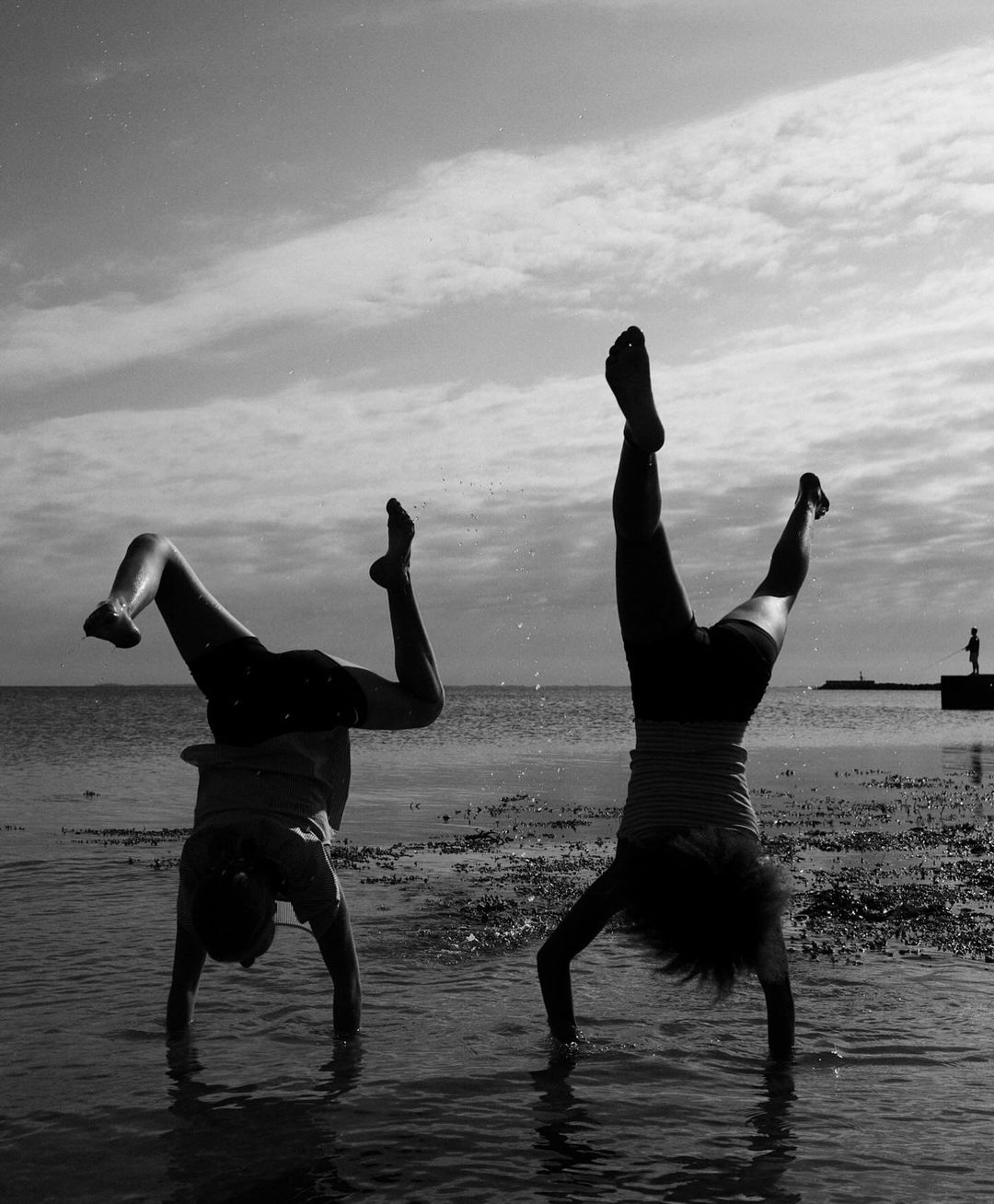 Two people doing handstands in shallow water at the beach in black and white, with cloudy sky and distant structure.