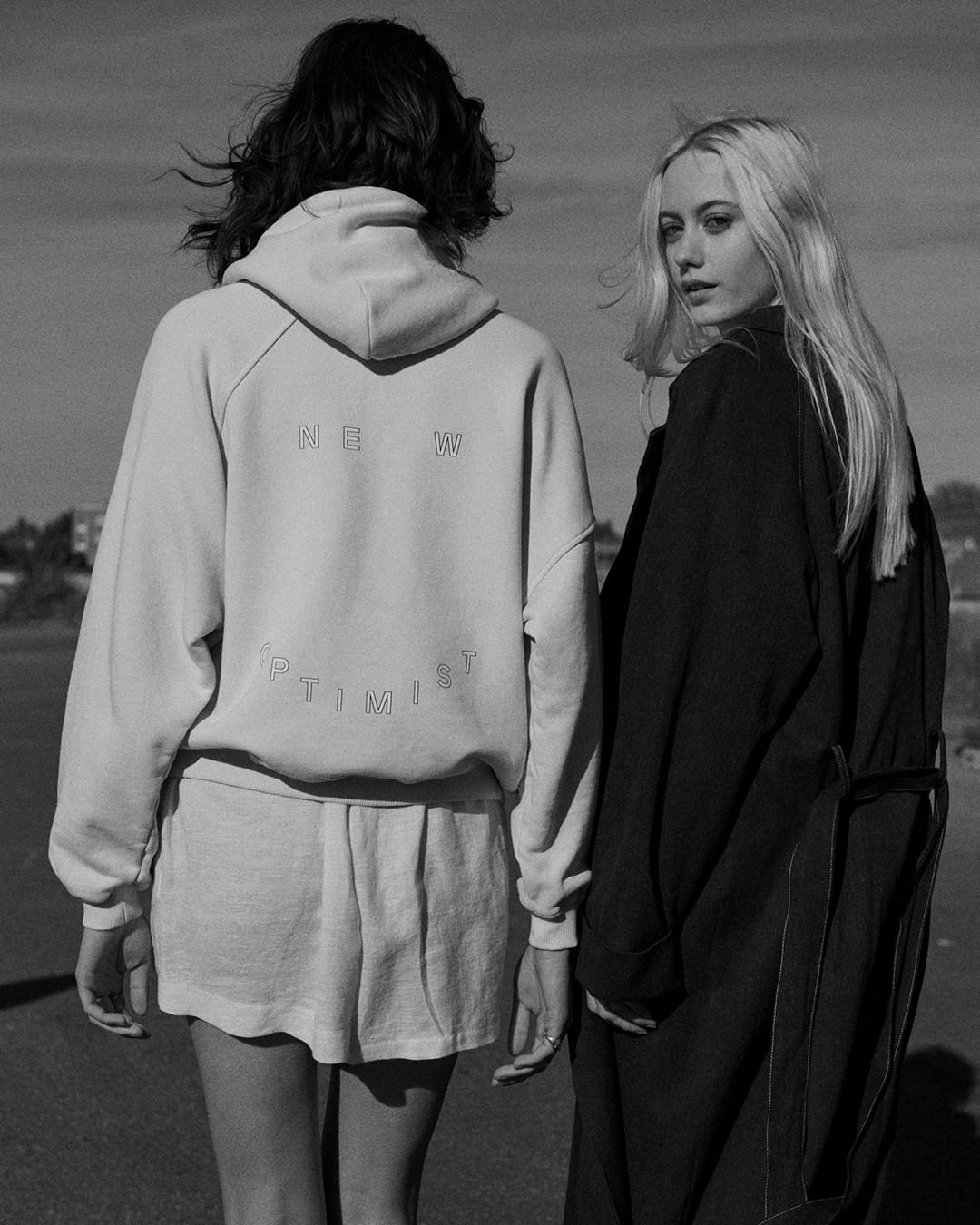 Two women stand outdoors on a sunny day, wearing a hoodie with text and a black coat, in a monochromatic image.