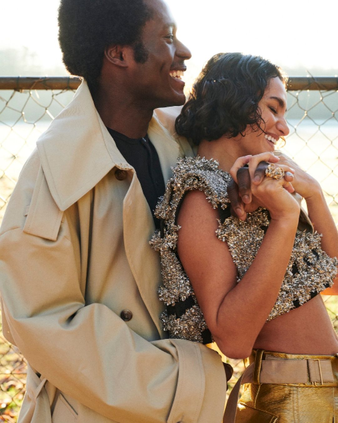 Smiling couple embraces in outdoor setting. Woman in metallic top and man in trench coat hold hands, backlit by natural light.