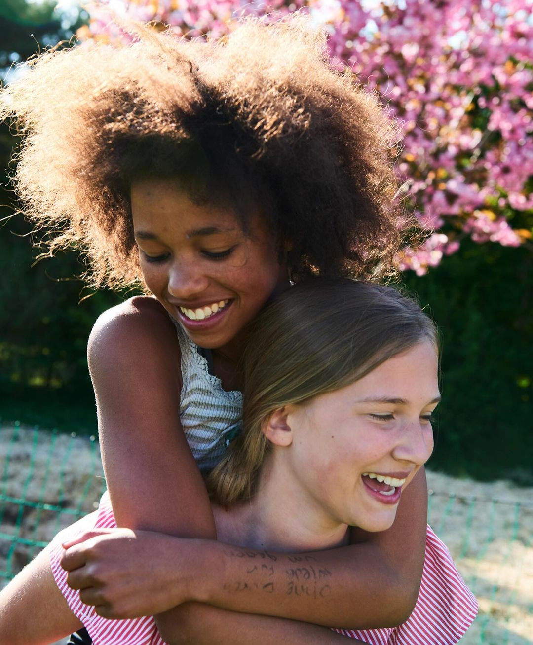 Two smiling girls, one with dark curly hair giving a piggyback ride to a light-skinned girl in outdoor light with pink flowers.