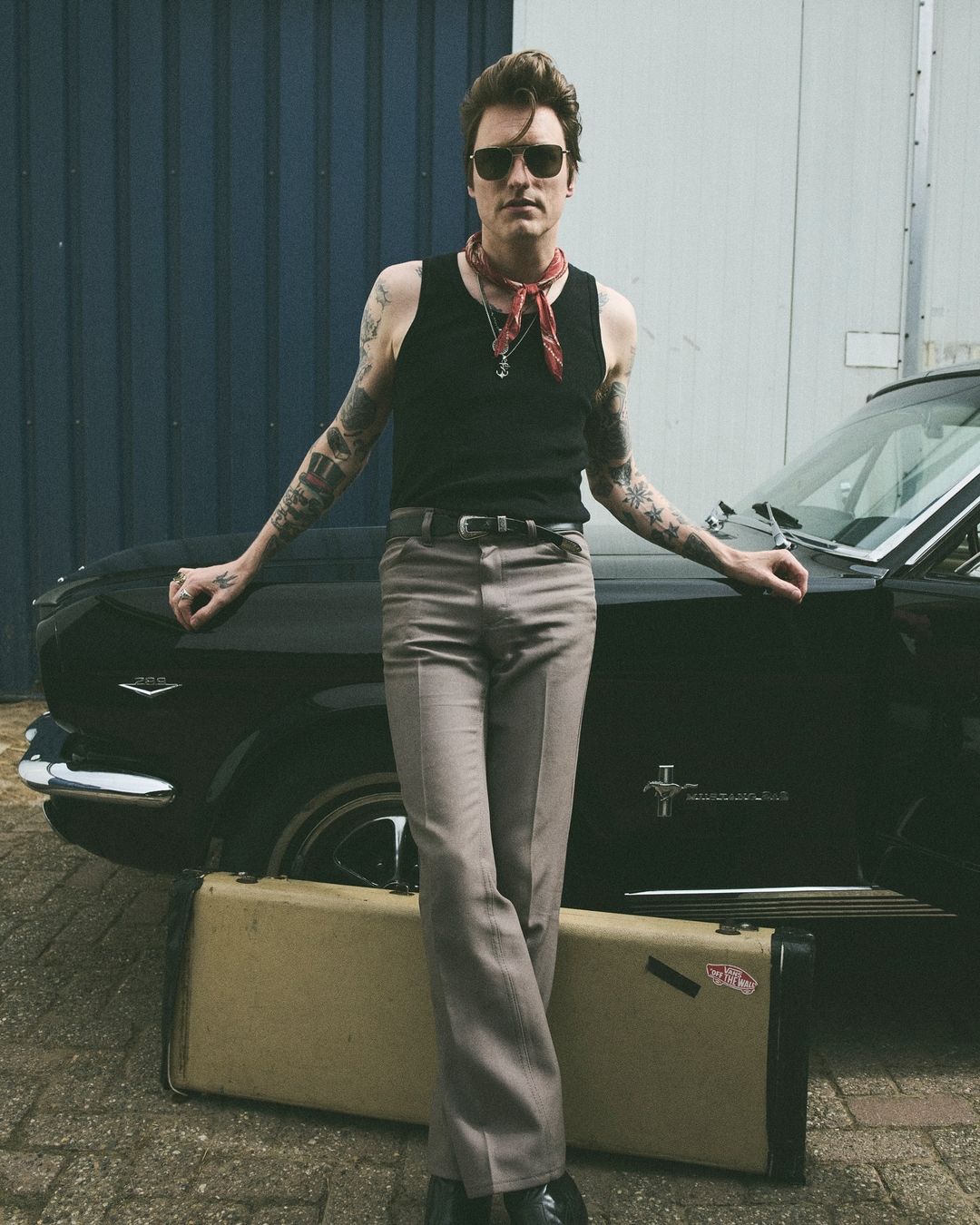 Man with sunglasses and tattoos poses by a black classic car with a beige hard case on a city street in daylight, wearing a bandana.