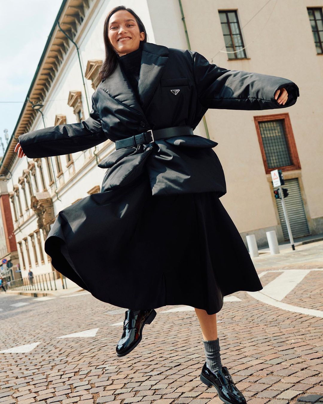 Fashion shot of an adult woman jumping on a cobblestone street wearing a black Prada coat, skirt, socks, and patent leather loafers.