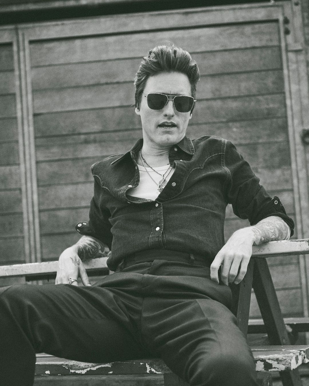 Black and white portrait of an adult male wearing sunglasses, denim shirt, and dark pants sitting outdoors with wood background.