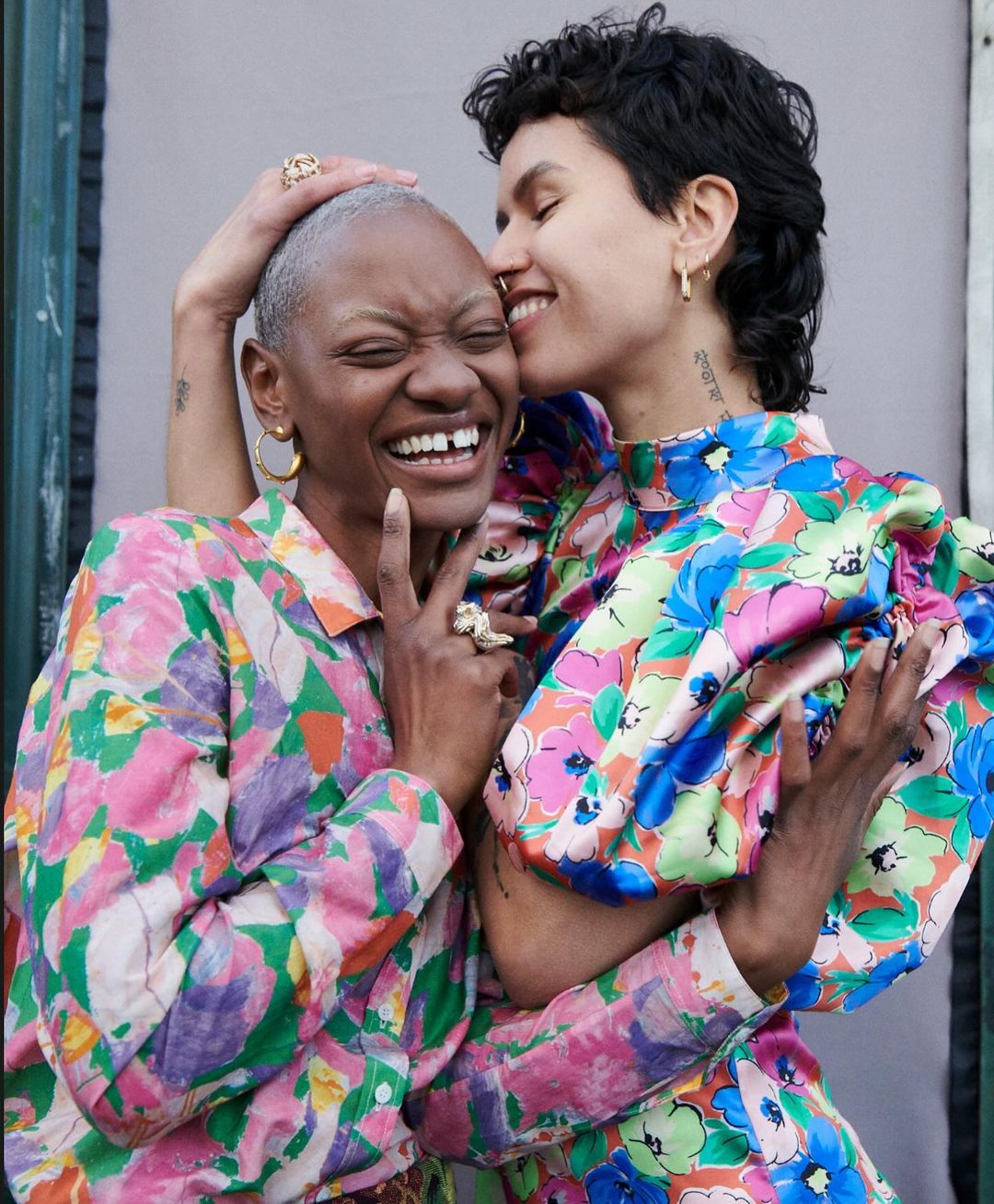 Two women in floral outfits embrace outdoors, one resting her head on the other's with a gray wall behind them.