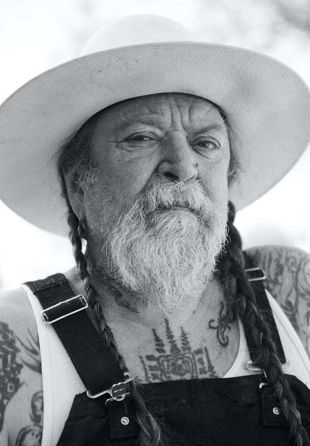 Close-up portrait of an elderly man with a long beard and braids, wearing a hat and overalls, showcasing tattoos on his chest and arms.