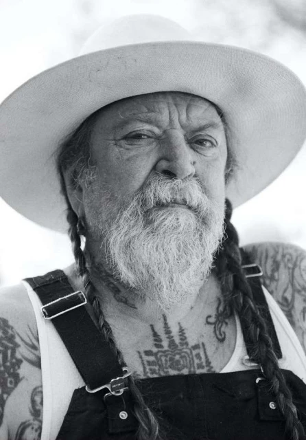 Close-up portrait of an elderly man with a long beard and braids, wearing a hat and overalls, showcasing tattoos on his chest and arms.