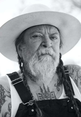 Close-up portrait of an elderly man with a long beard and braids, wearing a hat and overalls, showcasing tattoos on his chest and arms.