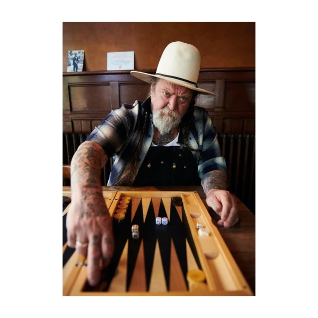 Man with a beard and cowboy hat playing backgammon at a wooden table in a warm-toned interior, focusing on the game and dice.