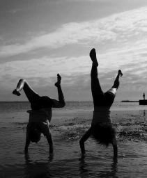 Two people doing handstands in shallow water at a beach under an overcast sky