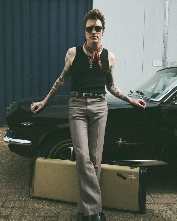 Man wearing black tank top and sunglasses leaning against a black classic car with a guitar case at his feet.