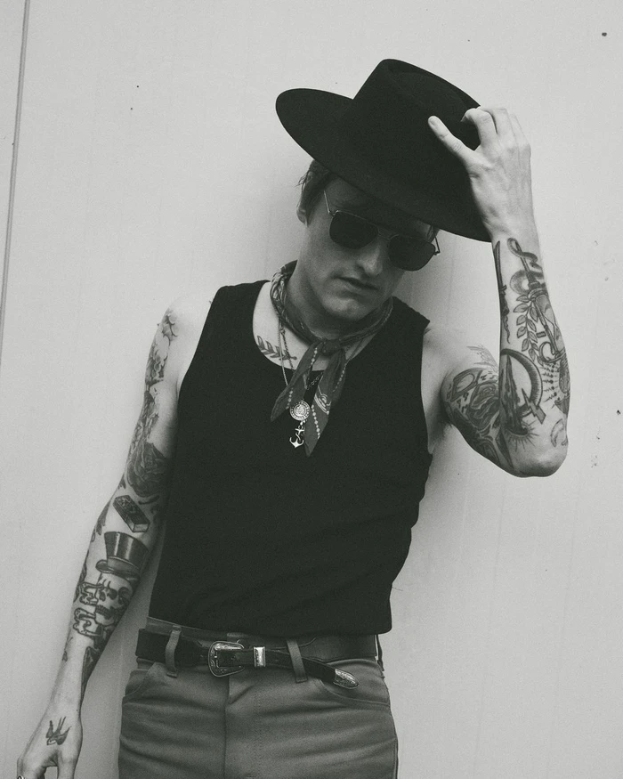 Man wearing a black fedora hat, sunglasses, tank top, and neck scarf with heavily tattooed arms, photographed in black and white.