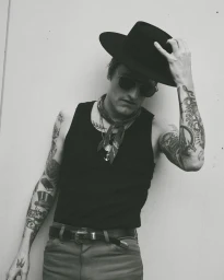 Man wearing a black fedora hat, sunglasses, tank top, and neck scarf with heavily tattooed arms, photographed in black and white.