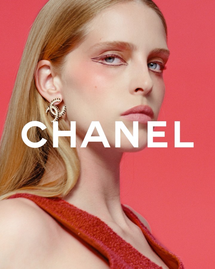 Close-up shot of a model showcasing beauty and fashion, featuring distinct eye makeup, jewelry, and a coral-colored backdrop with CHANEL text overlay.