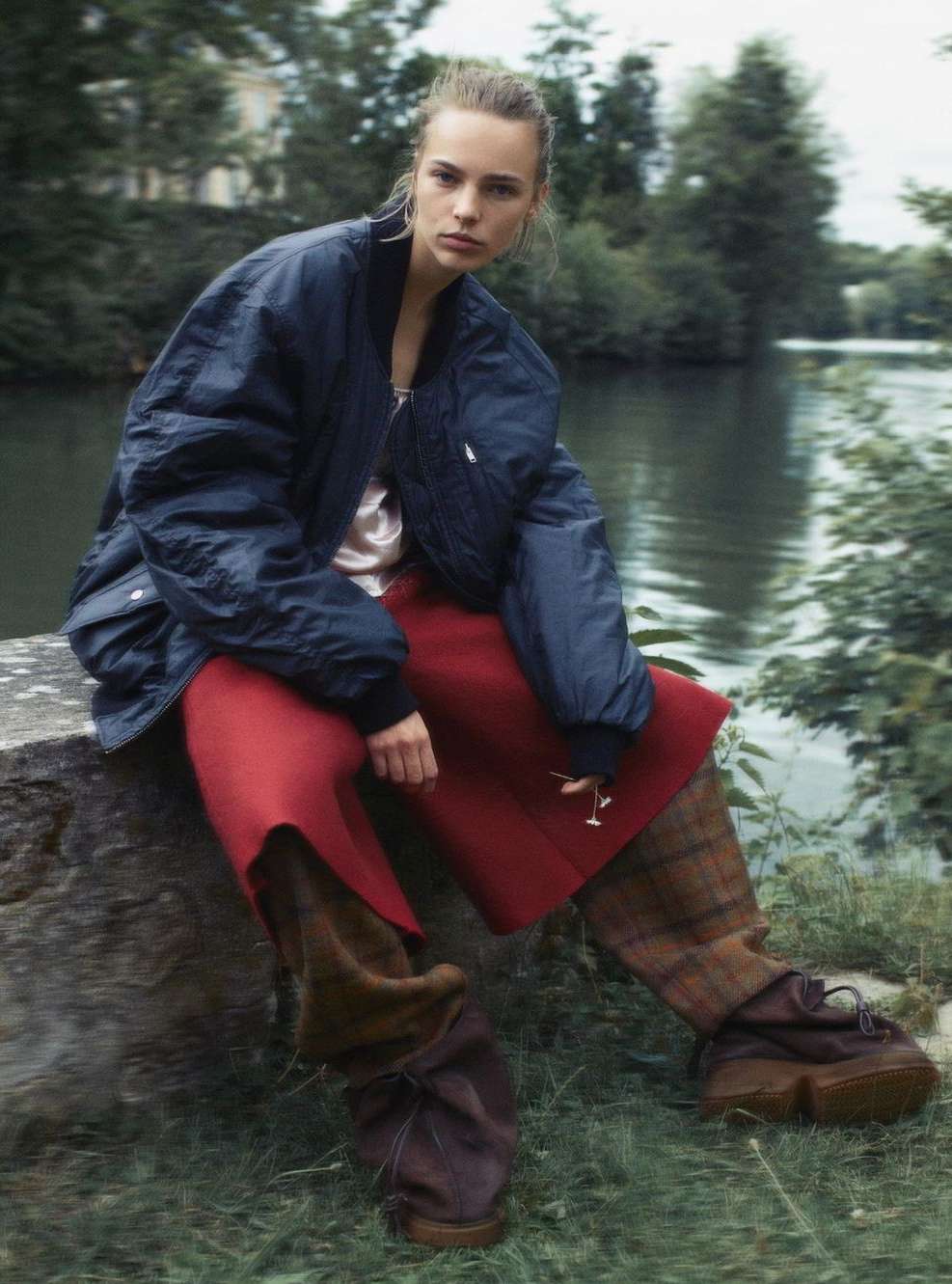 A woman with fair skin is sitting outdoors on a rock wearing a dark bomber jacket, rust-colored pants, and unique brown leather shoes.