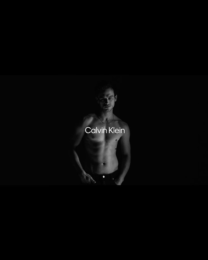 Black and white studio shot of a muscular adult male posing shirtless with Calvin Klein logo visible on torso against dark background.
