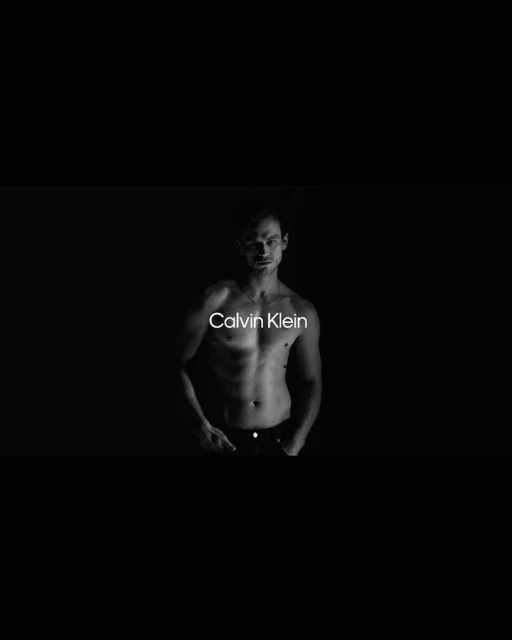 Black and white studio shot of a muscular adult male posing shirtless with Calvin Klein logo visible on torso against dark background.