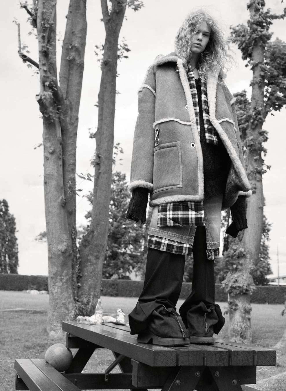 Woman with curly hair stands on a wooden picnic table in a shearling coat, black pants, and gloves in a monochrome outdoor shot.