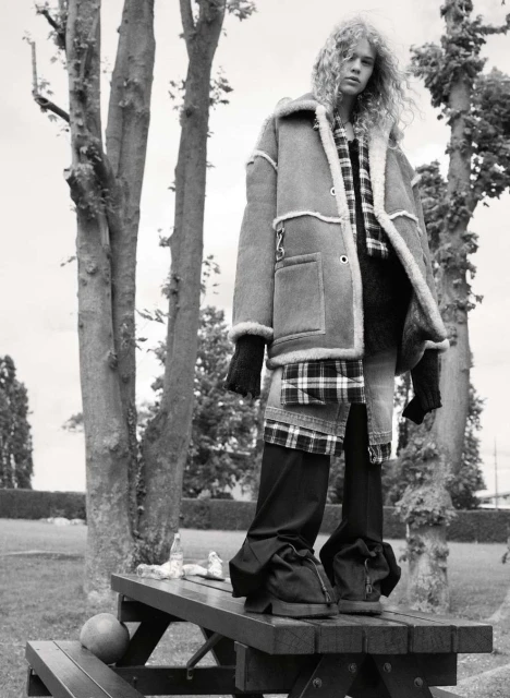 Woman with curly hair stands on a wooden picnic table in a shearling coat, black pants, and gloves in a monochrome outdoor shot.